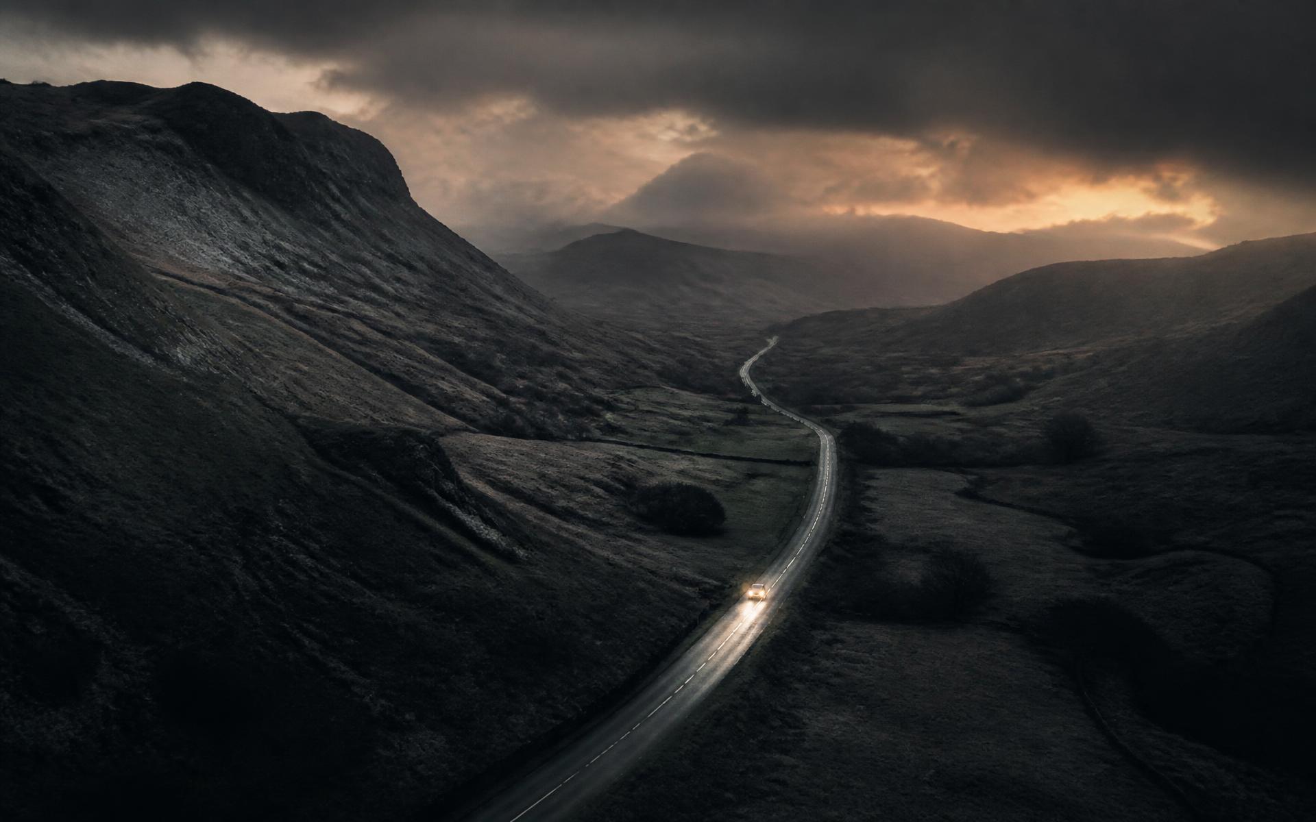 A road cutting through dramatic Cumbrian fells at dusk, a single vehicle heading out